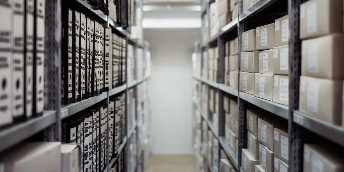 Lungo corridoio d'archivio con scaffali metallici grigi pieni di faldoni bianchi e neri identici fino al soffitto, etichette sfocate, il corridoio che si perde in una luce pallida