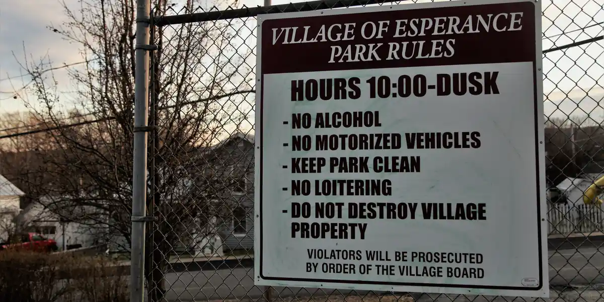 Cartello bianco fissato a una recinzione metallica con la scritta VILLAGE OF ESPERANCE PARK RULES, HOURS 10:00-DUSK e cinque divieti elencati uno sotto l'altro, chiusi dalla riga VIOLATORS WILL BE PROSECUTED BY ORDER OF THE VILLAGE BOARD