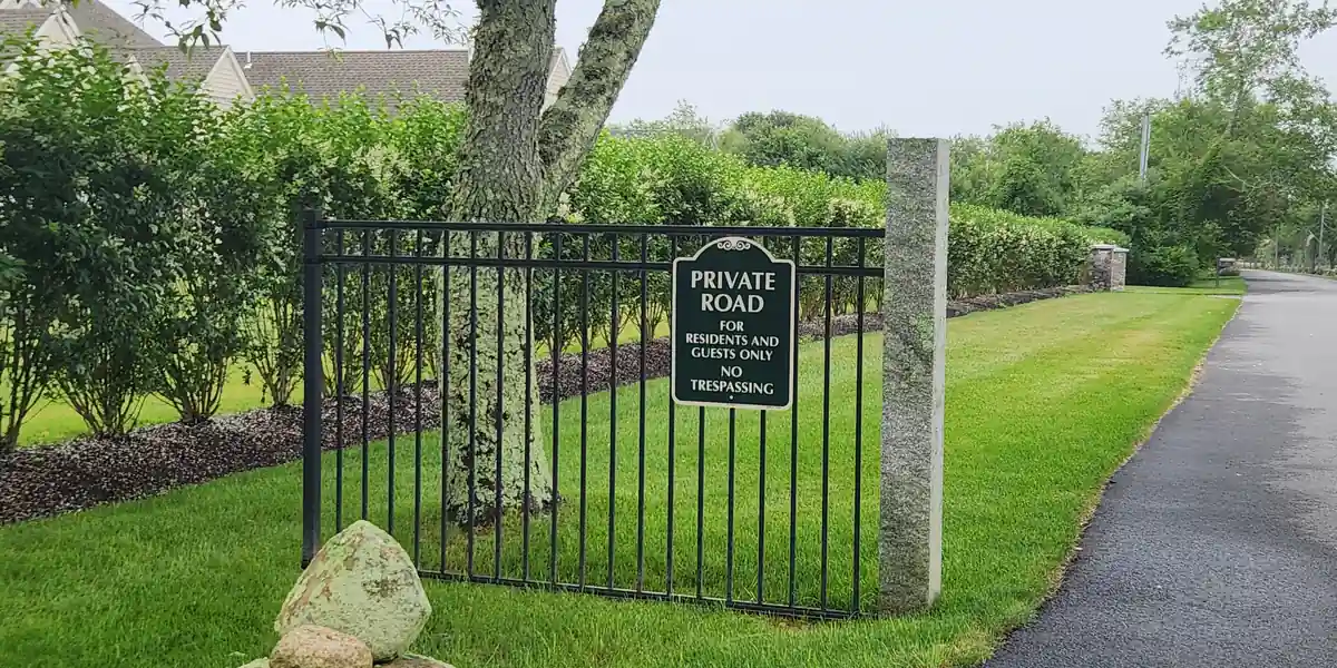 Breve cancellata in ferro battuto con una targa ovale che riporta PRIVATE ROAD, FOR RESIDENTS AND GUESTS ONLY, NO TRESPASSING, mentre subito accanto il vialetto asfaltato prosegue libero senza alcuna barriera