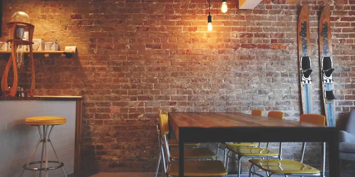Empty café interior: bare brick wall, two lit filament bulbs, a long wooden table with six chairs, a bar stool, nobody at any of them
