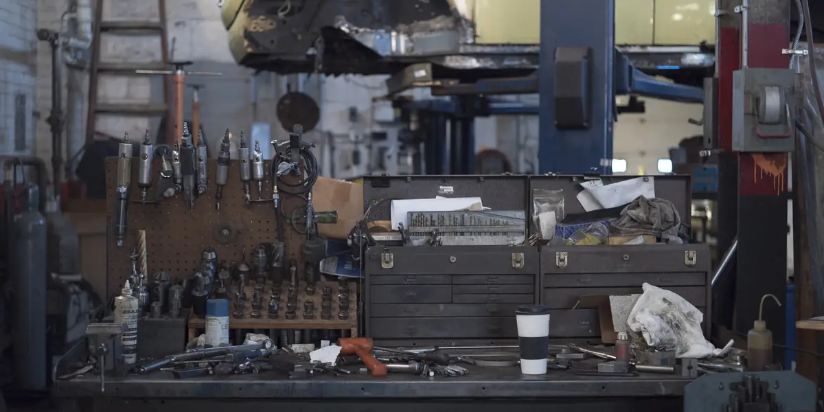 Mechanic's workshop: pegboard of air tools, open steel toolbox, drill index, a car body raised on a blue two-post lift behind the bench